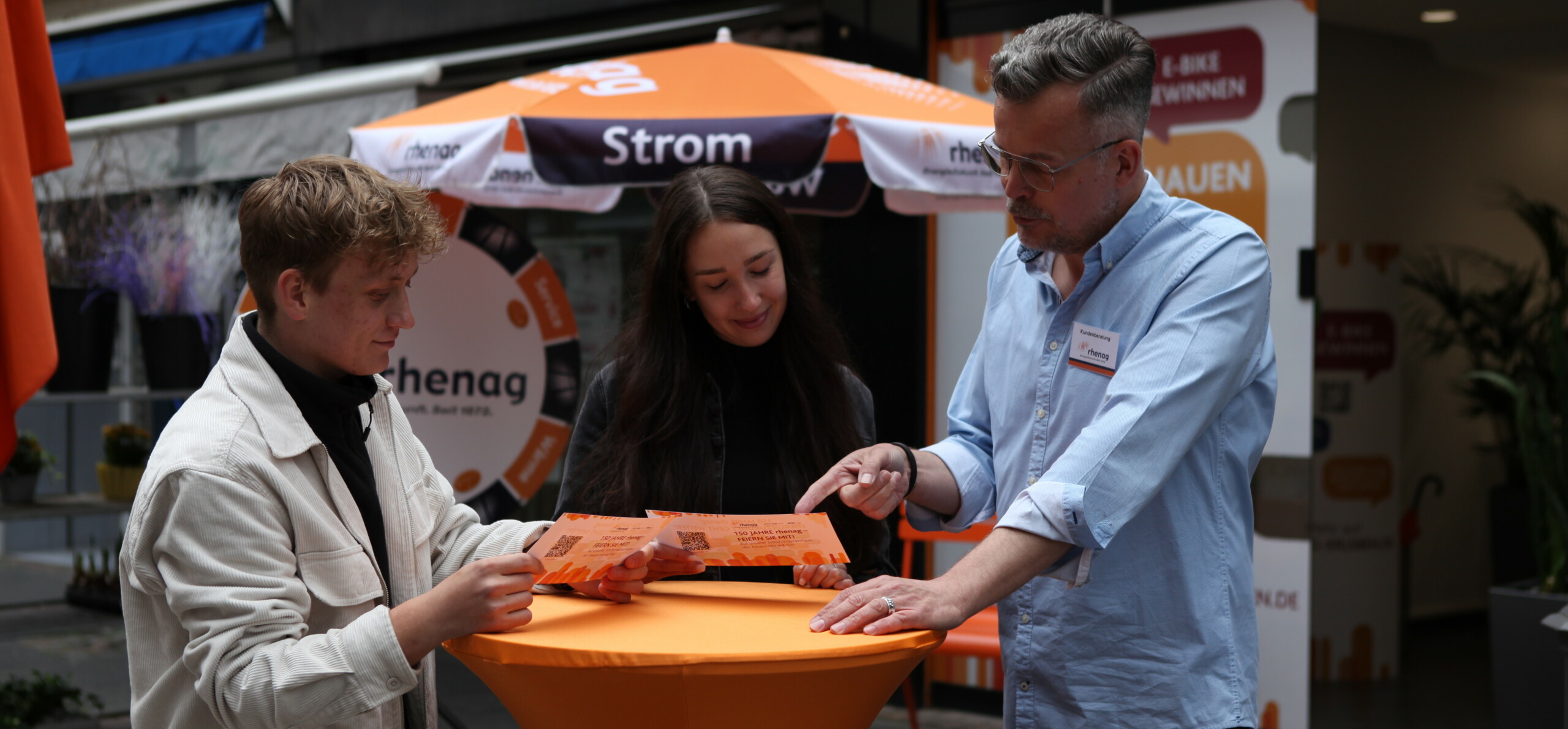 rhenag Mitarbeiter stehen an einem Stehtisch vor dem rhenag Shop und schauen sich Flyer an.
