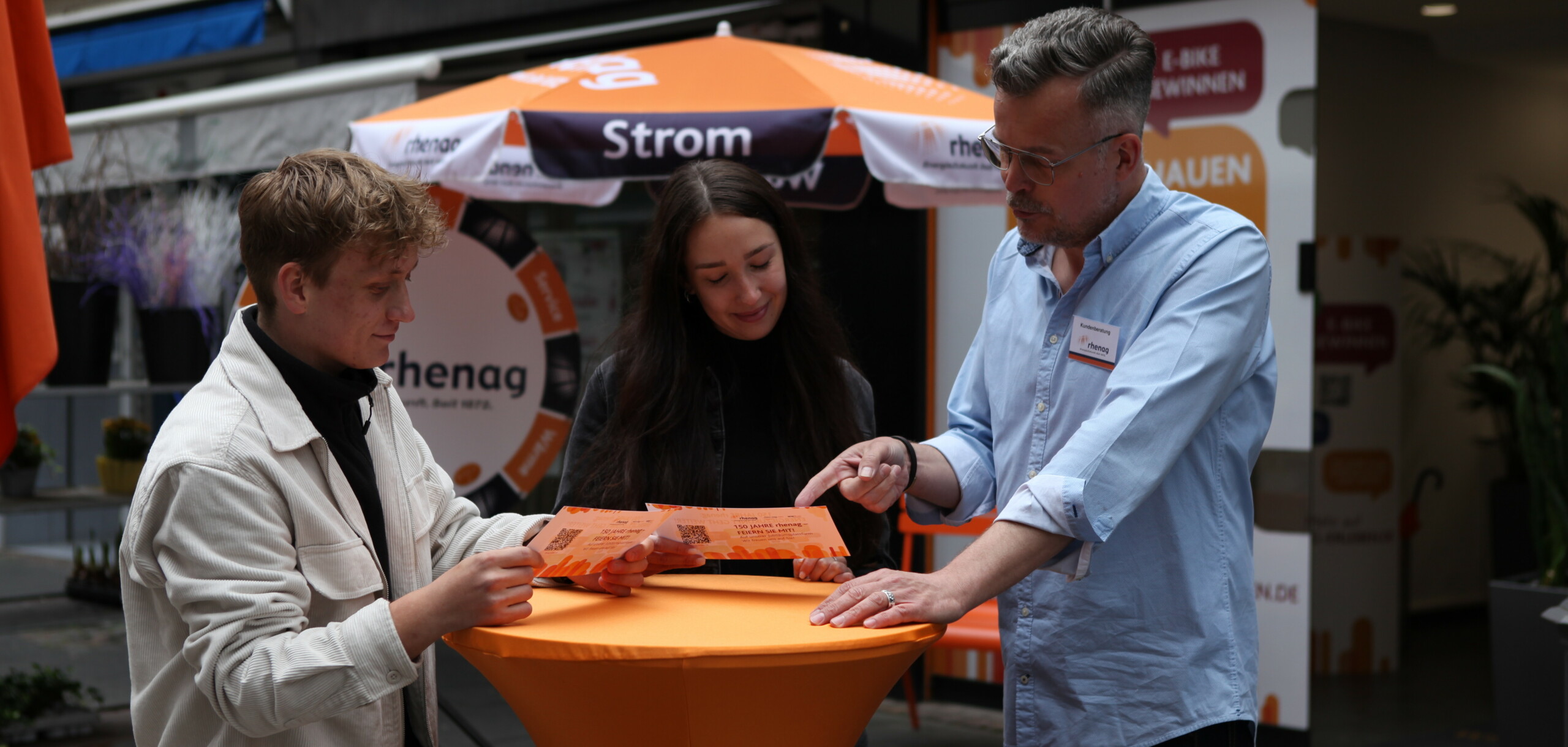Shop Siegburg Beratung rhenag Mitarbeiter stehen an einem Stehtisch vor dem rhenag Shop und schauen sich Flyer an.